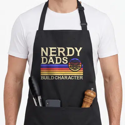 Nerdy gaming apron - Retro text NERDY DADS BUILD CHARACTER over blue red yellow orange stripes and d20 planet — shown on a black cotton canvas apron against a white background
