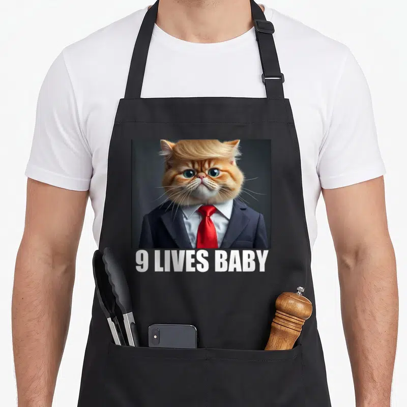 Trump cat apron - Orange cat in blue suit with red tie and white 9 LIVES BABY text below — shown on a black cotton canvas apron against a white background