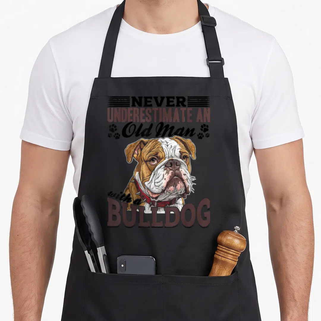 English top dog aprons - Cartoon bulldog portrait with spiked collar and text NEVER UNDERESTIMATE AN Old Man — shown on a black cotton canvas apron against a white background