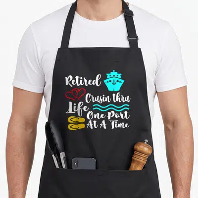 Cruisin retirement apron - Stacked white text reading Retired Crusin thru Life One Port At A Time with cyan ship, red — shown on a black cotton canvas apron against a white background