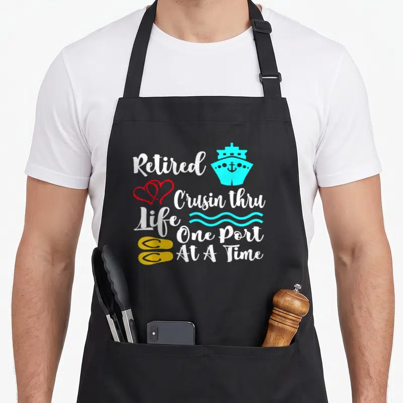 Cruisin retirement apron - Stacked white text reading Retired Crusin thru Life One Port At A Time with cyan ship, red — shown on a black cotton canvas apron against a white background