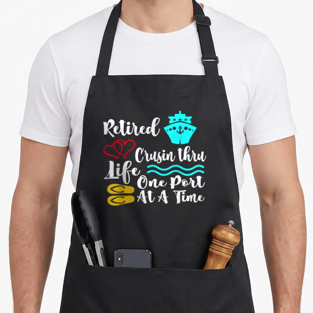 Cruisin retirement apron - Stacked white text reading Retired Crusin thru Life One Port At A Time with cyan ship, red — shown on a black cotton canvas apron against a white background