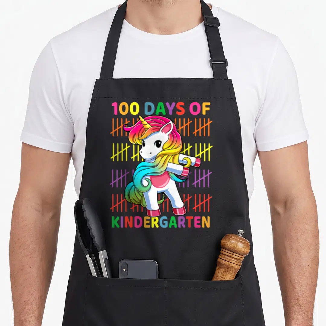 Kindergarten school unicorn aprons - Flossing cartoon unicorn with 100 Days of Kindergarten text and rainbow tally marks — shown on a black cotton canvas apron against a white background