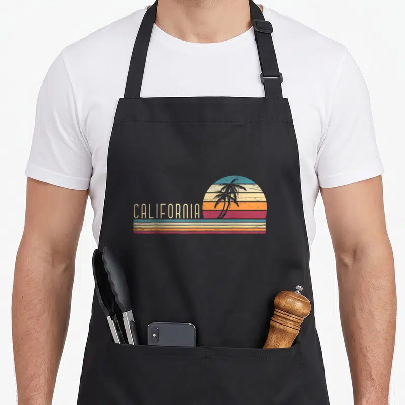 Vacation tree apron - Retro California sunset with palm tree silhouette and striped colors — shown on a black cotton canvas apron against a white background