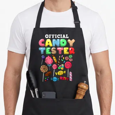 Candy tester halloween apron - OFFICIAL CANDY TESTER text above colorful lollipops cotton candy and wrapped sweets — shown on a black cotton canvas apron against a white background