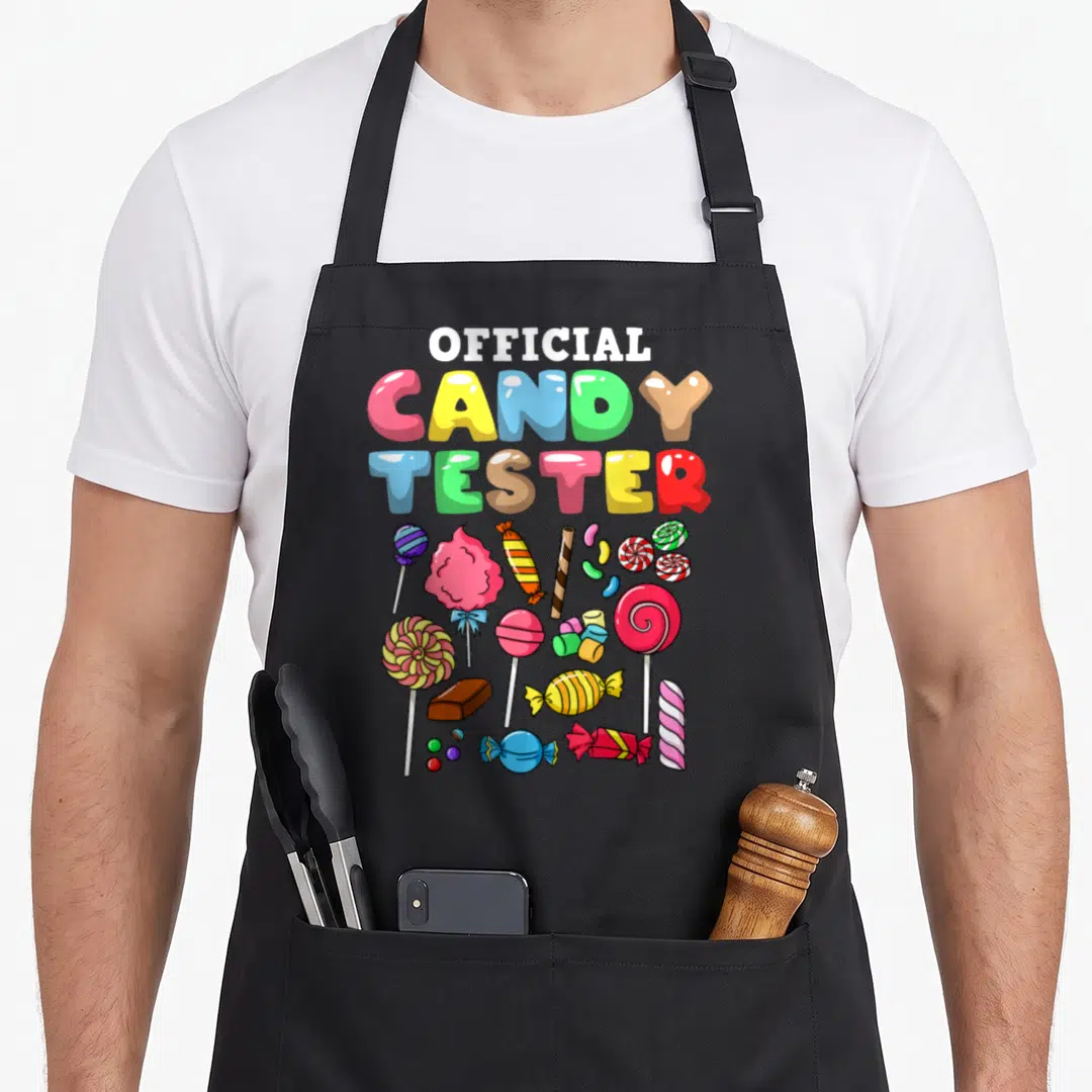 Candy tester halloween apron - OFFICIAL CANDY TESTER text above colorful lollipops cotton candy and wrapped sweets — shown on a black cotton canvas apron against a white background
