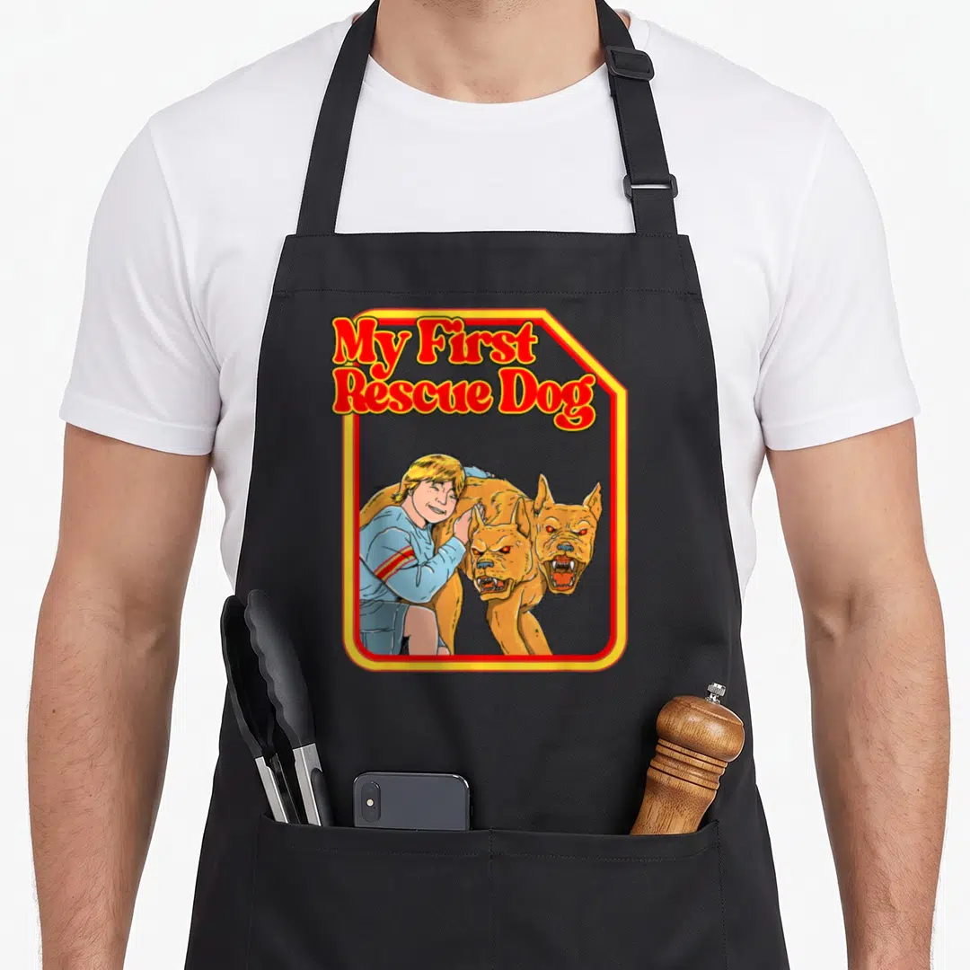 Dark grill rescue apron - Child hugging two-headed tan dog under My First Rescue Dog text in red and yellow border — shown on a black cotton canvas apron against a white background