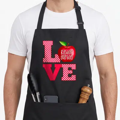 Funny kindergarten teacher apron - Red polka dot LOVE text with apple containing Kinder garten script — shown on a black cotton canvas apron against a white background