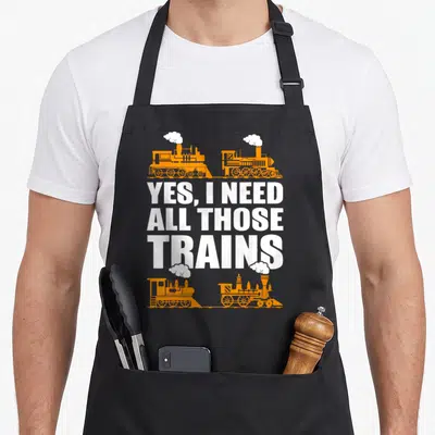Collector railroad train apron - Four orange steam trains surrounding white text reading Yes I need all those trains — shown on a black cotton canvas apron against a white background