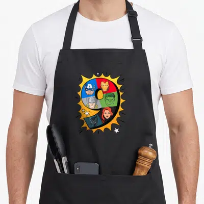 Heroe 9th birthday apron - Number 9 divided into colored segments with superhero faces inside a yellow comic burst — shown on a black cotton canvas apron against a white background