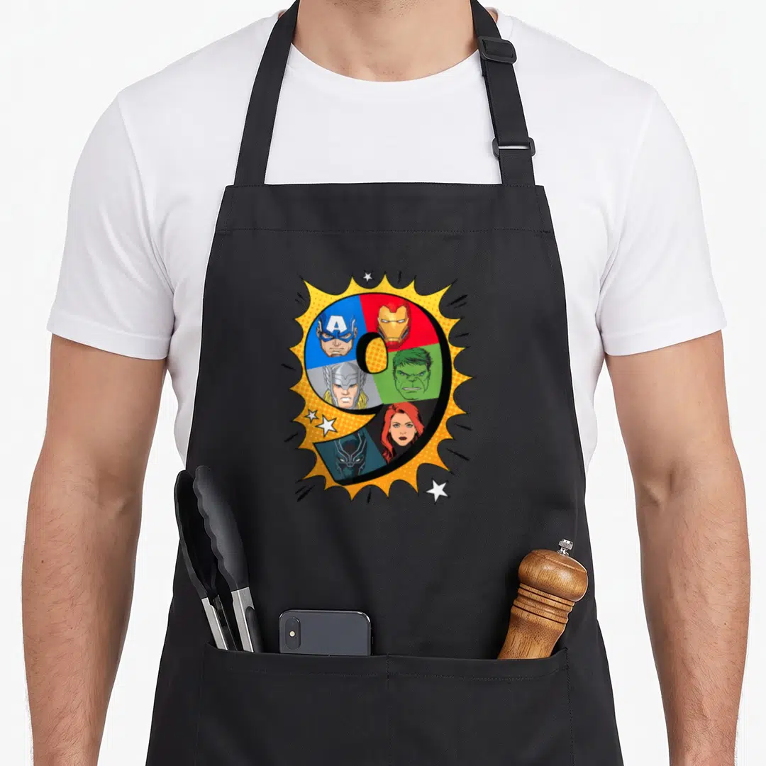 Heroe 9th birthday apron - Number 9 divided into colored segments with superhero faces inside a yellow comic burst — shown on a black cotton canvas apron against a white background