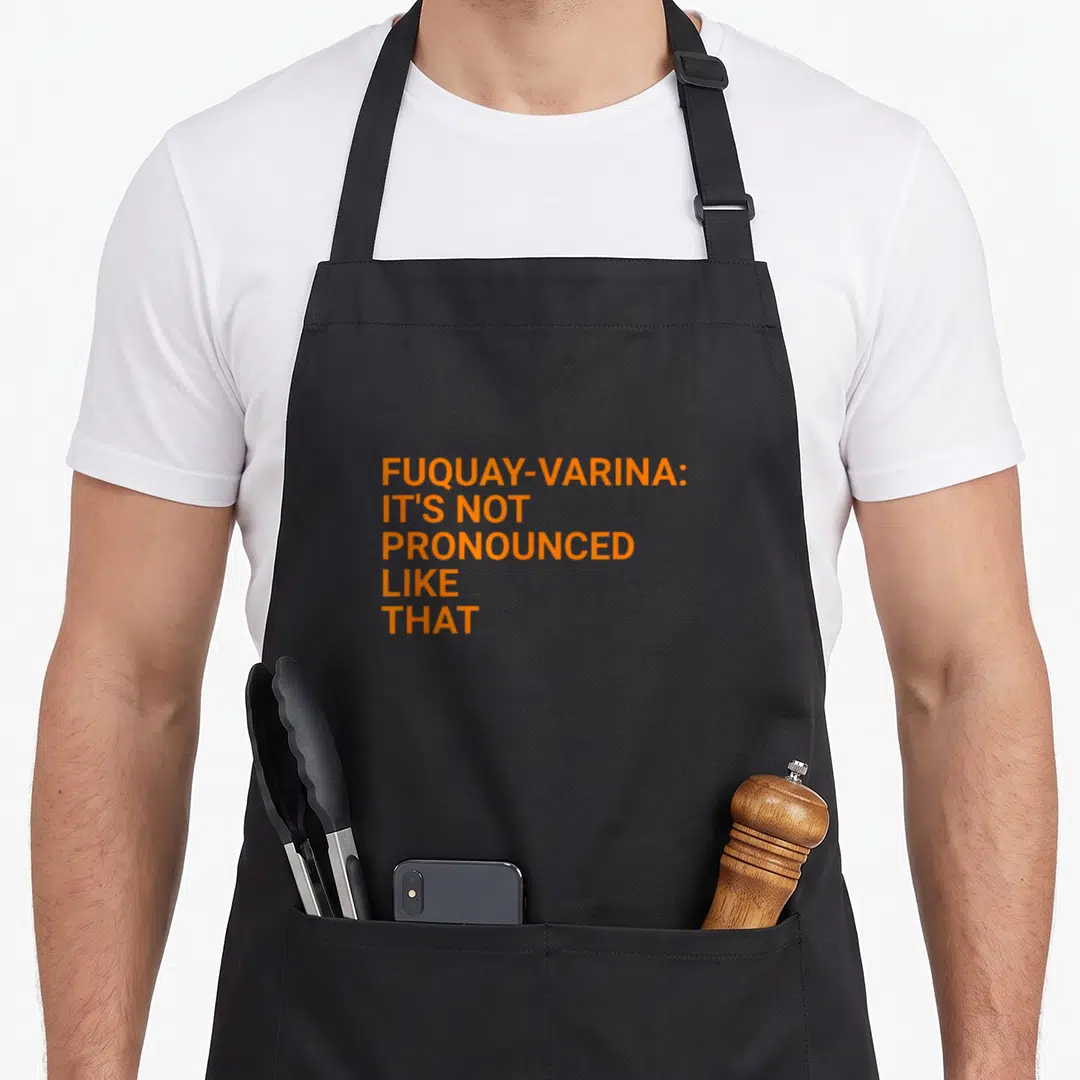 Fuquay pride apron - Orange stacked text reading FUQUAY-VARINA IT'S NOT PRONOUNCED LIKE THAT — shown on a black cotton canvas apron against a white background