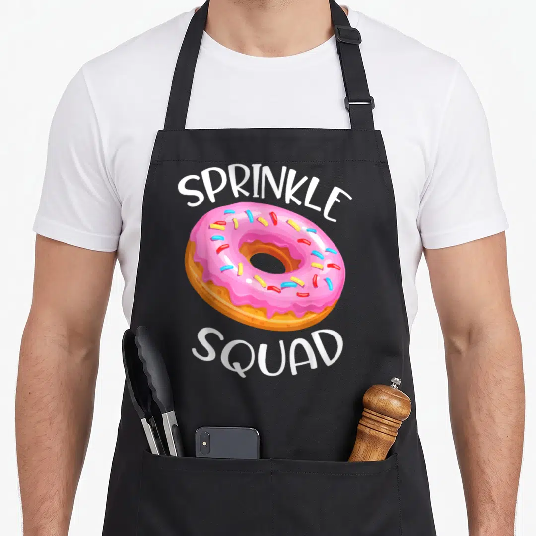 Donut sprinkle apron - Pink frosted donut with colorful sprinkles between two arched lines of white text — shown on a black cotton canvas apron against a white background