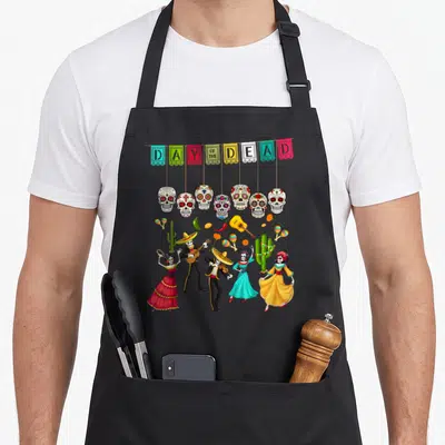 Muerto mexican apron - Day of the Dead banners, sugar skulls, and dancing skeleton musicians in red yellow blue green — shown on a black cotton canvas apron against a white background