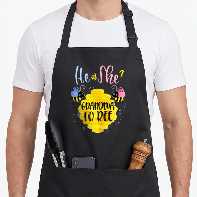 She bee apron - Cartoon bees flanking yellow honeycomb with He or She text — shown on a black cotton canvas apron against a white background