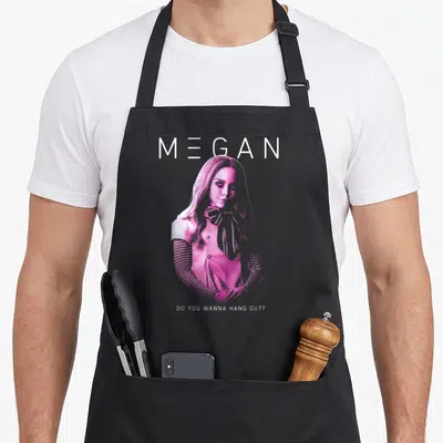 M3gan nbc apron - Pink woman illustration with MEGAN text and DO YOU WANNA HANG OUT question — shown on a black cotton canvas apron against a white background