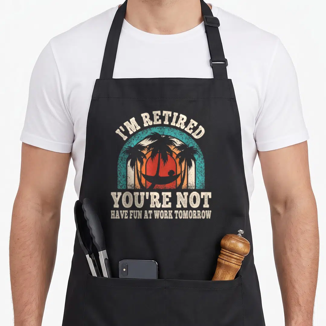 Retire funny retirement aprons - I'M RETIRED YOU'RE NOT text over teal orange sunset beach scene with palm trees — shown on a black cotton canvas apron against a white background