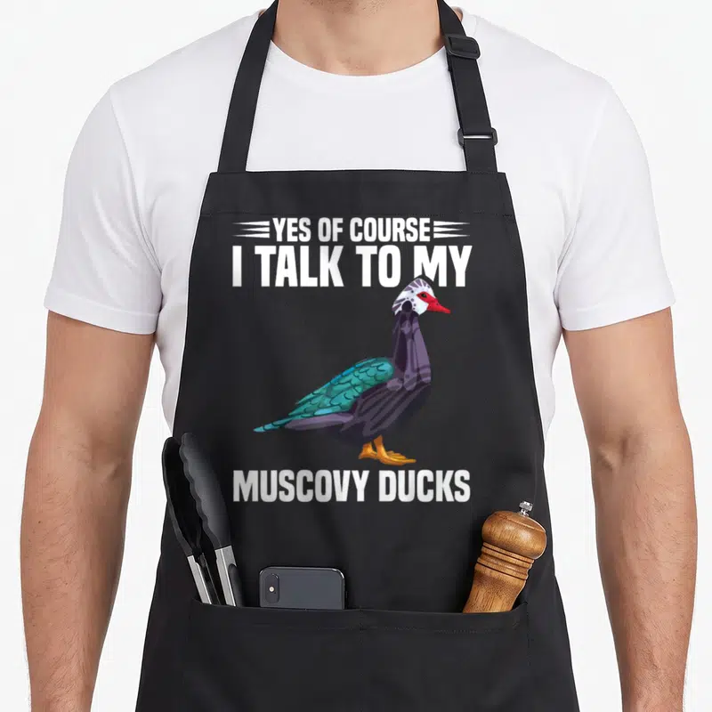Necklace cotton duck apron - Cartoon Muscovy duck with red beak and teal wings framed by white text on black fabric — shown on a black cotton canvas apron against a white background