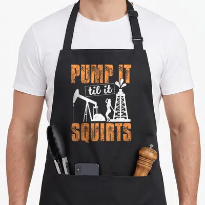 Pump oil apron - PUMP IT til it SQUIRTS text with pumpjack and oil rig silhouettes in orange and white — shown on a black cotton canvas apron against a white background