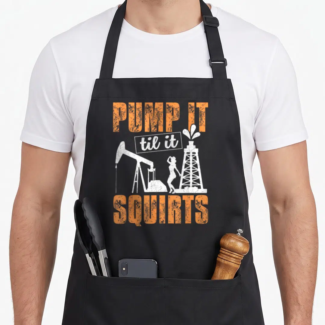 Pump oil apron - PUMP IT til it SQUIRTS text with pumpjack and oil rig silhouettes in orange and white — shown on a black cotton canvas apron against a white background