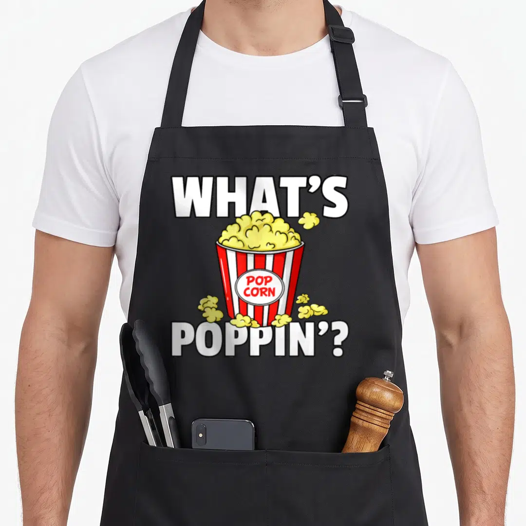 Poppin popcorn apron - Cartoon red white striped popcorn bucket with yellow kernels and text WHAT'S POP CORN POPPIN' — shown on a black cotton canvas apron against a white background