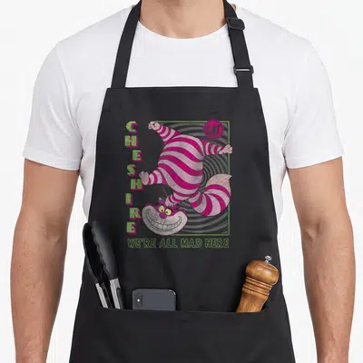 Cheshire cat alice wonderland apron - Upside down pink striped cat in grey spiral frame with We're All Mad Here text — shown on a black cotton canvas apron against a white background