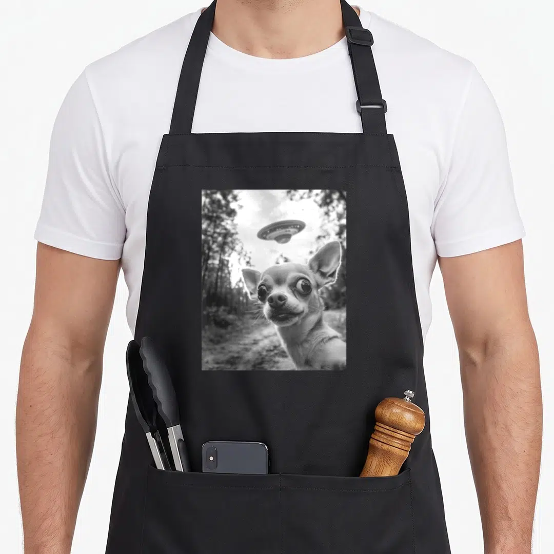 Chihuahua meme apron - Black and white photo of chihuahua face with UFO hovering above trees — shown on a black cotton canvas apron against a white background