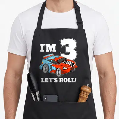 Toddler car apron - Cartoon race car with I'M 3 LET'S ROLL text in blue red orange — shown on a black cotton canvas apron against a white background