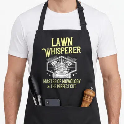 Lawn landscaper problems apron - Lawn Whisperer text and lawn mower graphic between crossed rakes in a hexagon frame — shown on a black cotton canvas apron against a white background
