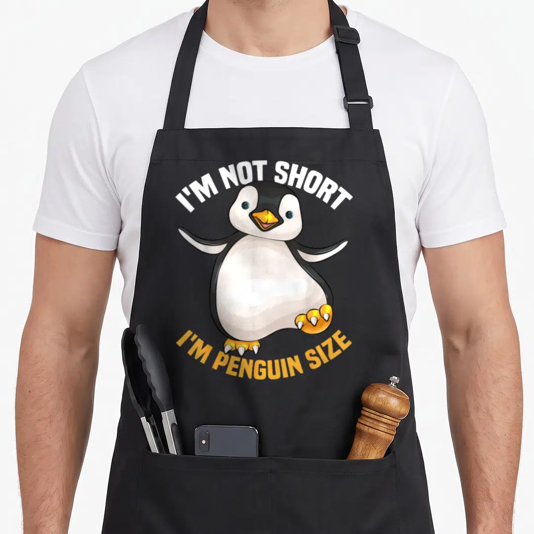 Fan penguin aprons - Cartoon penguin with arched text I'M NOT SHORT I'M PENGUIN SIZE in white and orange — shown on a black cotton canvas apron against a white background
