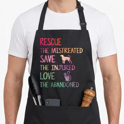 Pet animal rescue apron - Stacked rainbow text reading Rescue Save Love with dog and cat silhouettes — shown on a black cotton canvas apron against a white background