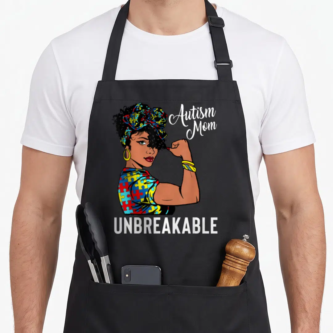 Afro mother's apron - Cartoon woman flexing arm with autism mom text and puzzle piece clothing in red blue yellow — shown on a black cotton canvas apron against a white background