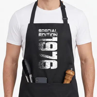 Cute special birthday apron - White distressed text reading Special Edition 1976 stacked vertically on black fabric — shown on a black cotton canvas apron against a white background