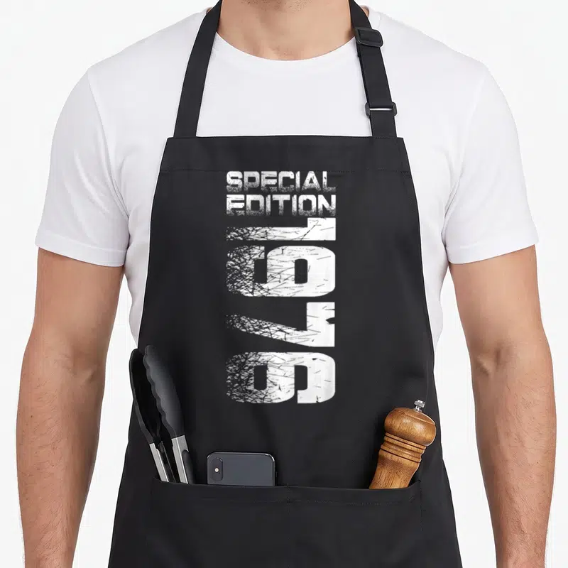 Cute special birthday apron - White distressed text reading Special Edition 1976 stacked vertically on black fabric — shown on a black cotton canvas apron against a white background
