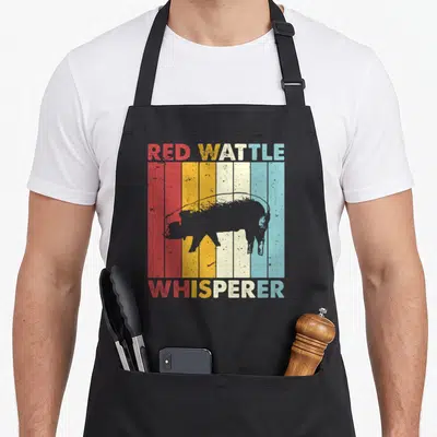 Breeder farmer apron - Retro Red Wattle Whisperer text with black pig silhouette over red yellow white blue stripes — shown on a black cotton canvas apron against a white background