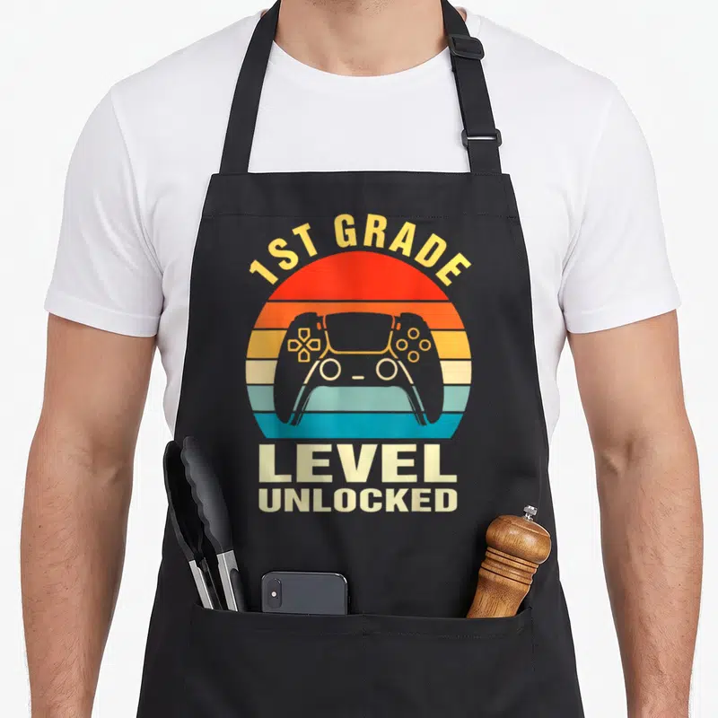 Grade unlocked first apron - Retro striped sun with centered game controller and 1st Grade Level Unlocked text — shown on a black cotton canvas apron against a white background