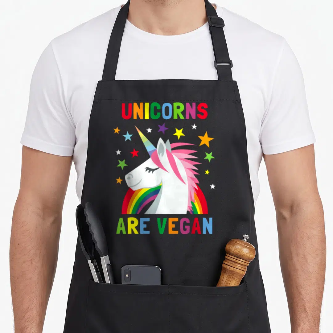 Unicorn plant based apron - Cartoon unicorn head with pink mane over rainbow surrounded by stars and text — shown on a black cotton canvas apron against a white background