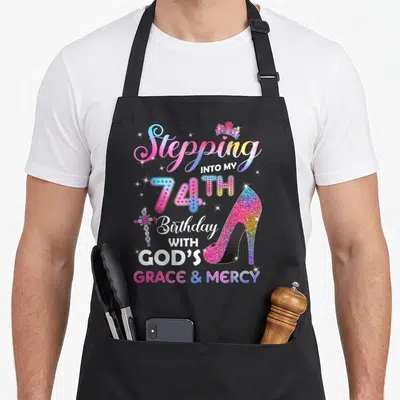 Stepping 74th birthday apron - Rainbow high heel shoe, jeweled cross, crown, and stacked birthday text — shown on a black cotton canvas apron against a white background