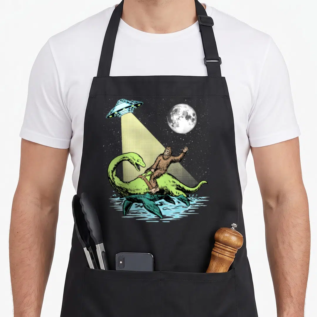 Nessie bigfoot apron - Cartoon bigfoot riding green plesiosaur under yellow ufo beam with moon — shown on a black cotton canvas apron against a white background