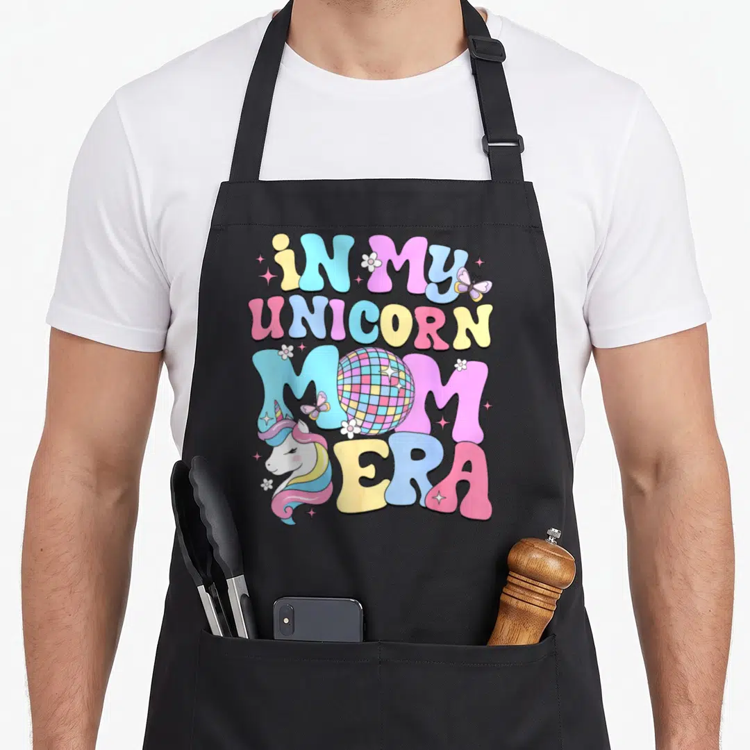 Unicorn mother's day aprons - Stacked text IN MY UNICORN MOM ERA with disco ball in MOM, unicorn head left of ERA — shown on a black cotton canvas apron against a white background