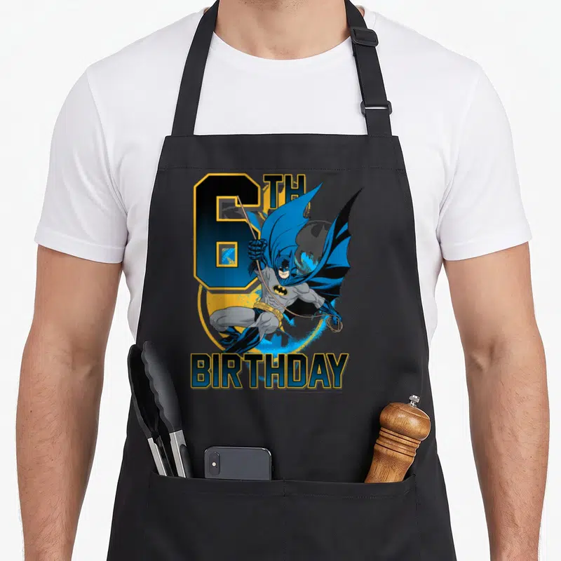 Action birthday apron - Leaping Batman graphic with 6TH BIRTHDAY text in blue yellow and grey — shown on a black cotton canvas apron against a white background