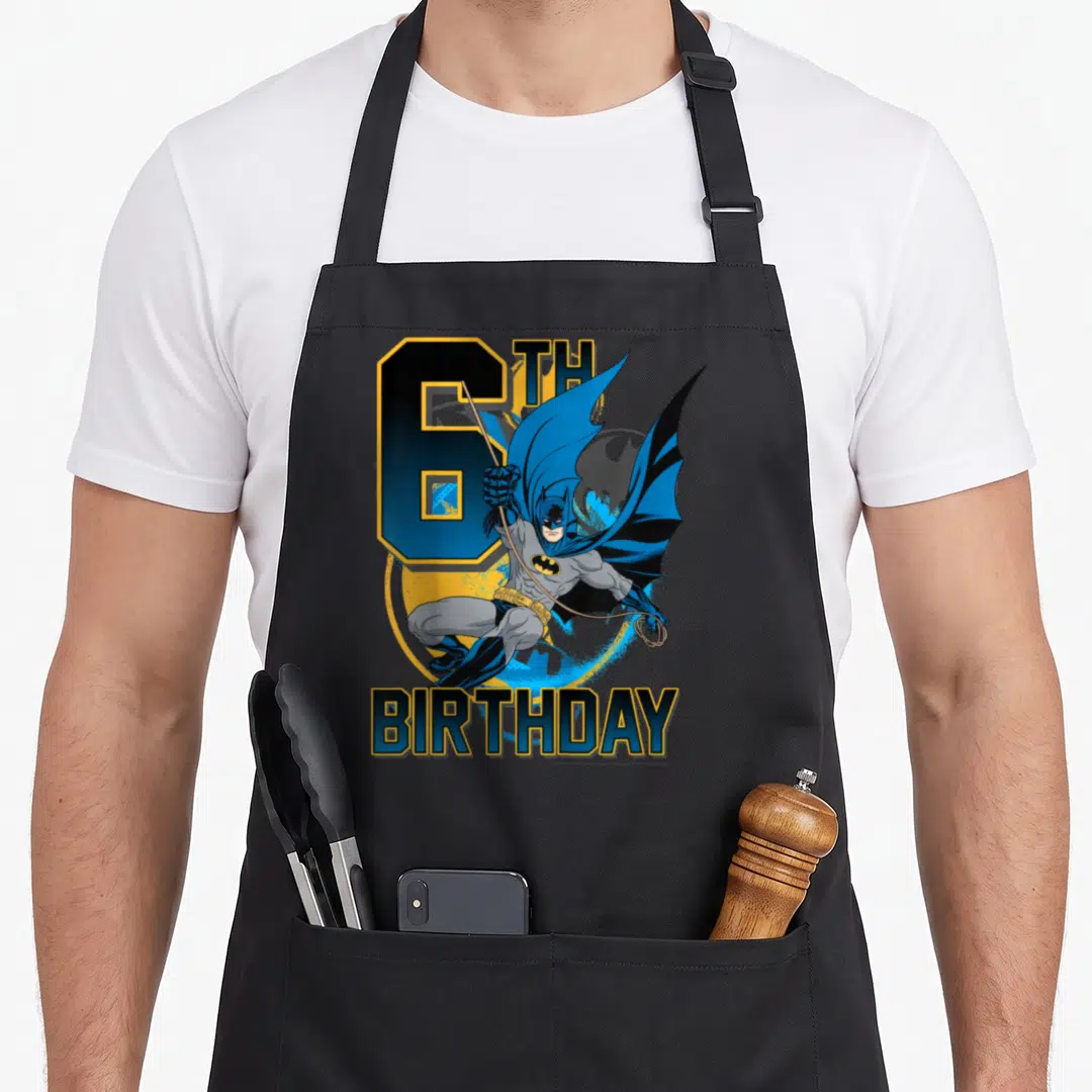 Action birthday apron - Leaping Batman graphic with 6TH BIRTHDAY text in blue yellow and grey — shown on a black cotton canvas apron against a white background