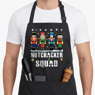 Squad nutcracker apron - Four cartoon nutcrackers on snow with NUTCRACKER SQUAD text and hanging ornament — shown on a black cotton canvas apron against a white background