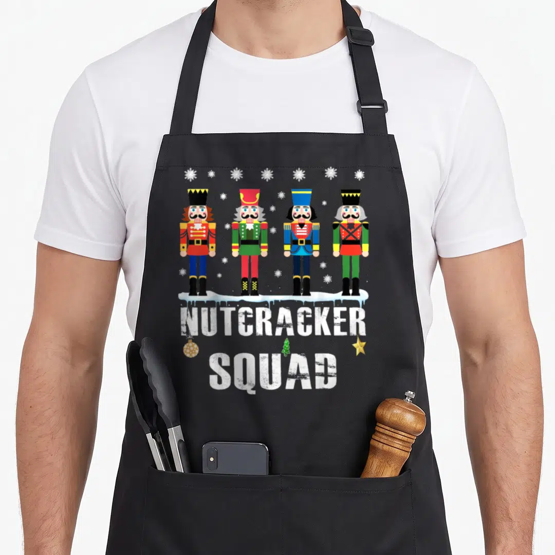 Squad nutcracker apron - Four cartoon nutcrackers on snow with NUTCRACKER SQUAD text and hanging ornament — shown on a black cotton canvas apron against a white background