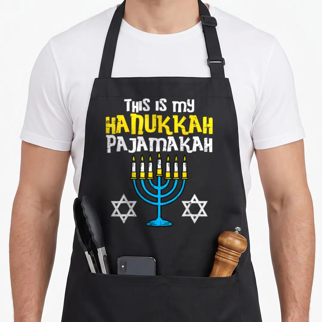 Pyjamas hanukkah apron - Stacked text THIS IS MY HANUKKAH PAJAMAKAH above blue menorah flanked by two white stars — shown on a black cotton canvas apron against a white background