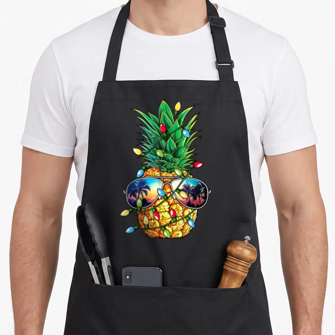 Pineapple christmas tree apron - Cartoon pineapple wearing sunglasses wrapped in red, blue, and yellow Christmas lights — shown on a black cotton canvas apron against a white background