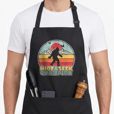 Sasquatch hide bigfoot apron - Retro bigfoot silhouette in a sunset circle with HIDE SEEK CHAMPION text below — shown on a black cotton canvas apron against a white background
