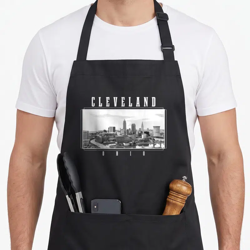Cleveland ohio pride apron - White CLEVELAND and O H I O text framing a grey scale city skyline photo on black fabric — shown on a black cotton canvas apron against a white background