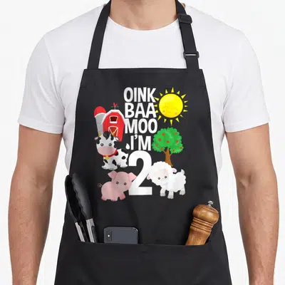 Oink farm apron - Oink Baa Moo I'm 2 text with cartoon cow pig sheep barn sun and apple tree — shown on a black cotton canvas apron against a white background