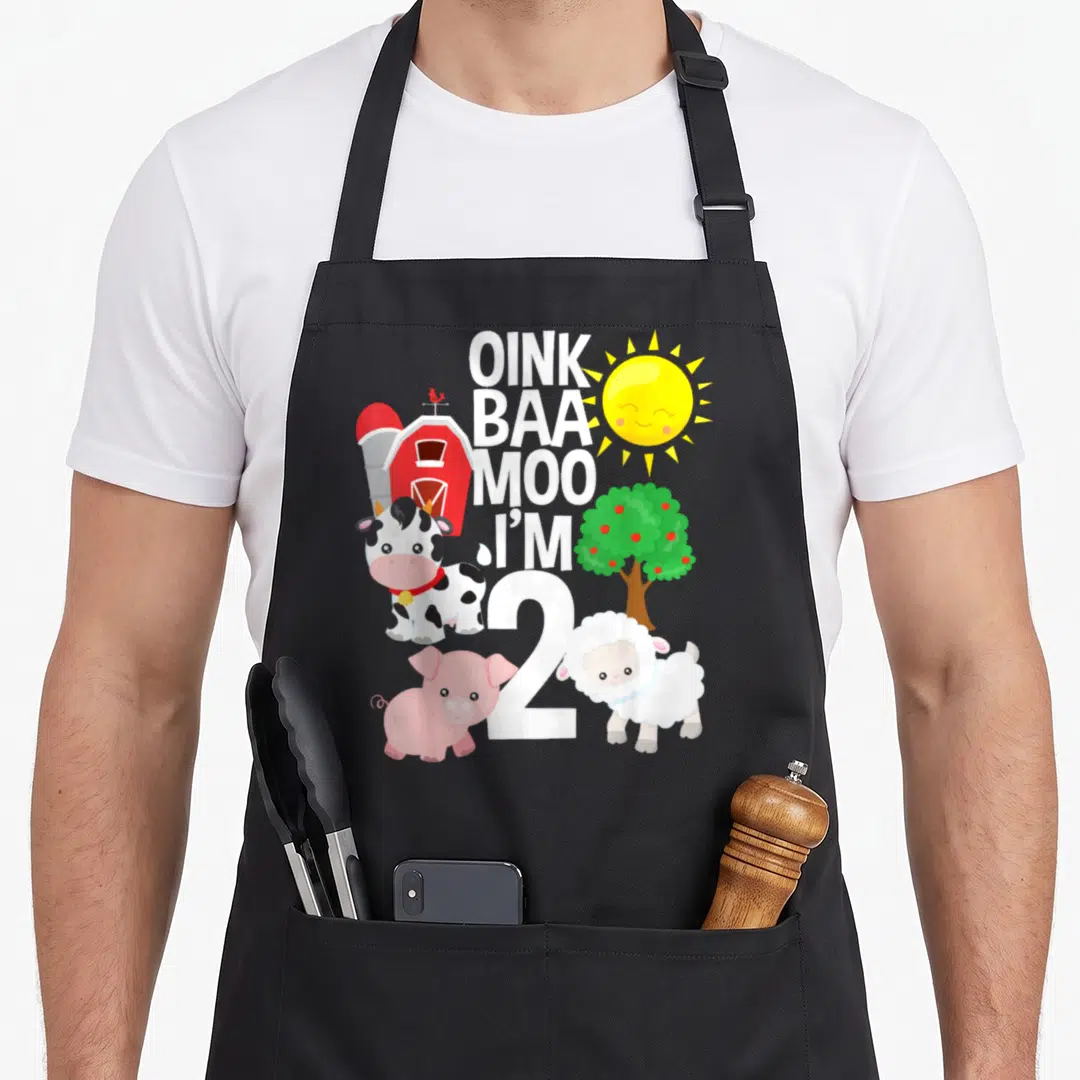 Oink farm apron - Oink Baa Moo I'm 2 text with cartoon cow pig sheep barn sun and apple tree — shown on a black cotton canvas apron against a white background